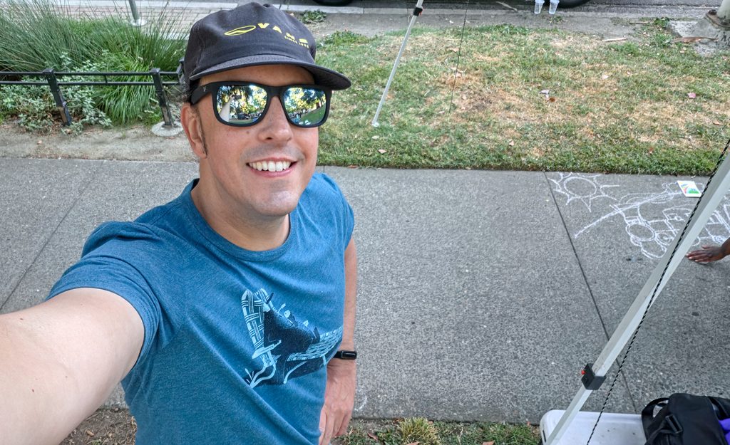 A selfie of me standing over a blank sidewalk square. I have a hat and sunglasses on, and I am smiling.