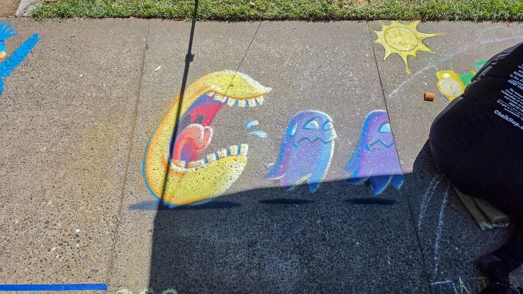 A photo showing the entire sidewalk square with the finished Pac-Man and ghosts in it. The sun is lighting part of the square and casting a shadow of my neighbor's tent over it.