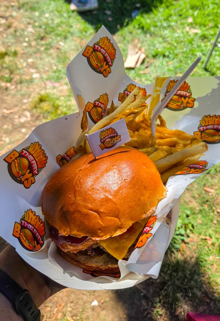 A photo of my hand holding a burger in a tray. The burger has a beef patty, cheese, onion rings, and bacon on it. Behind it is a bunch of fries covered in nacho cheese.