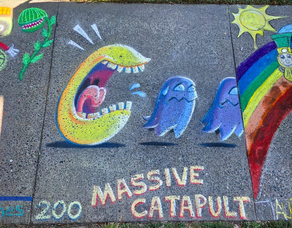 A photo of the finished artwork from a high angle. The finished art shows Pac-Man chasing two scared ghosts, with two drops of spit flying out of Pac-Man's mouth, shadows under all the subjects, and three lines emanating from Pac-Man's head. The lettering underneath the illustration (the square number and the words "Massive Catapult") is also blocked and finished.