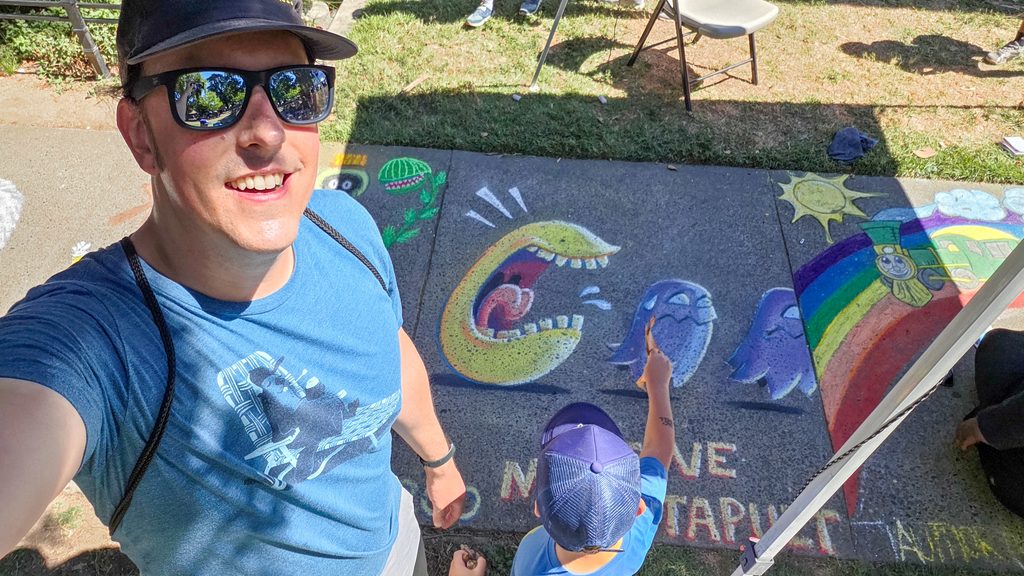 Another selfie of me standing over my finished artwork. It is a bit brighter than the first selfie, from the higher sun, and the finished artwork is on the sidewalk square behind me. My son is standing in front of the square, facing away from the camera, and holding a dried leaf over it.